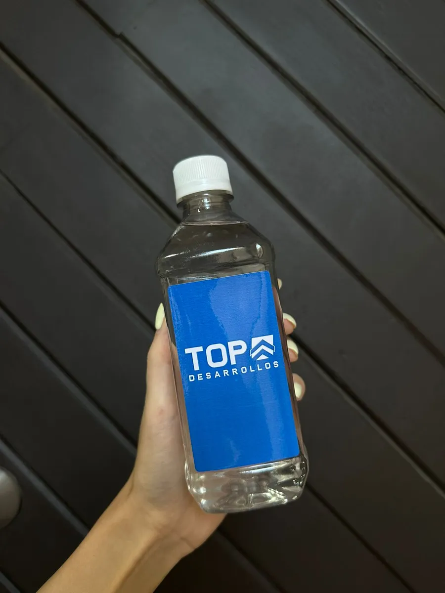 Mano sosteniendo una botella cuadrada de 500 ml con etiqueta vertical azul de una desarrolladora inmobiliaria